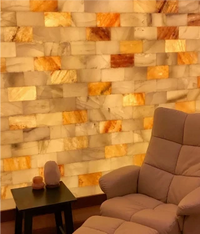 A warm-toned interior scene showing a beige upholstered lounge chair in front of a large wall made of rectangular polished stone tiles in cream, tan, and orange hues. A small dark side table with two small objects sits at the lower left.